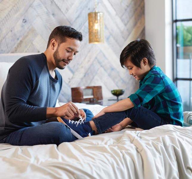 A father tying his young son's shoe laces while sitting on a bed.