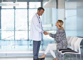 Female patient in waiting room meeting and shaking hand with male clinician in white lab coat.