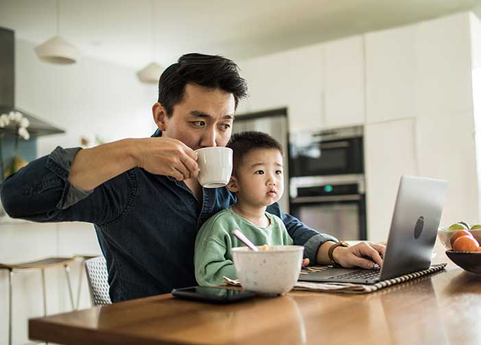Man at his work laptop drinking coffee with child on his lap in home kitchen