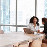 Two women sit across a wooden table in a bright office. One has a laptop, and they are engaged in a friendly conversation, conveying a collaborative tone.