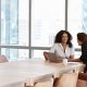 Two women sit across a wooden table in a bright office. One has a laptop, and they are engaged in a friendly conversation, conveying a collaborative tone.