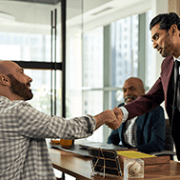 A man in a burgundy suit shakes hands with a seated, bearded man in a plaid shirt at a conference table. Two others men observing.