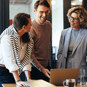 Four people stand around a table with a laptop, papers, and drinks, engaged in a discussion and smiling in a modern office setting
