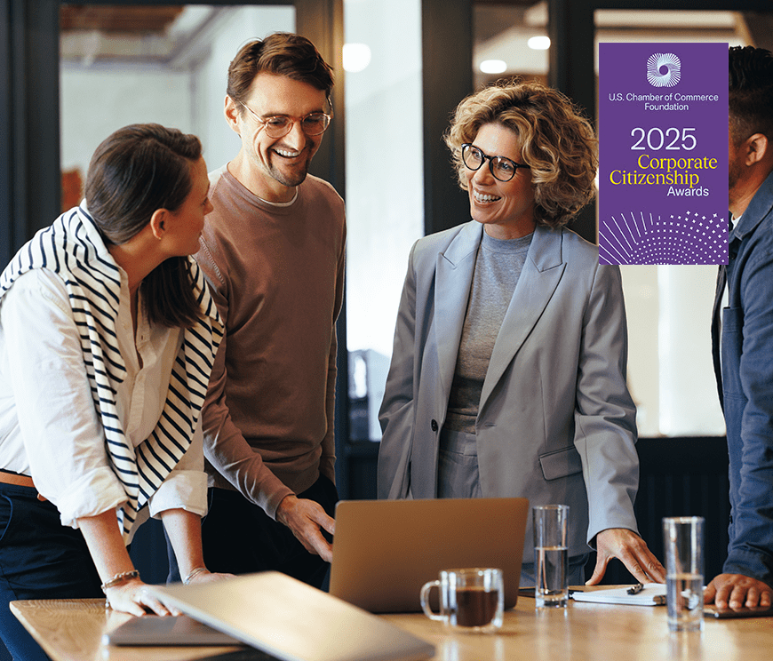 Four people stand around a table with a laptop, papers, and drinks, engaged in a discussion and smiling in a modern office setting