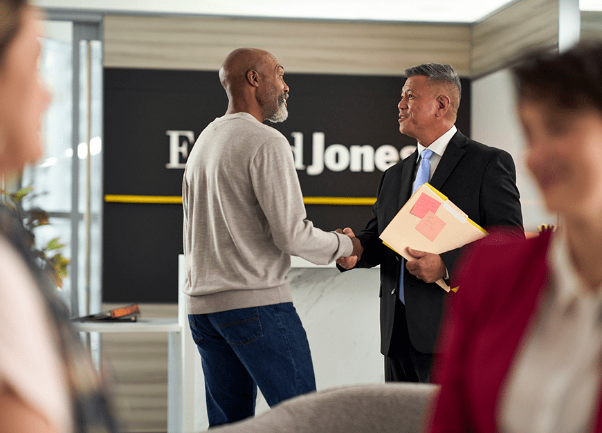 An office setting where two men shake hands, one in a suit holding documents, standing in front of a Edward Jones sign.