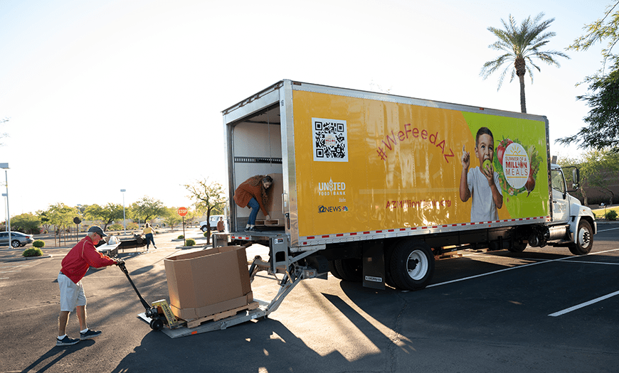 A man in a red shirt operates a pallet jack to load boxes onto a truck with a bright yellow "WeFeedAZ" banner featuring a smiling child.