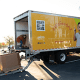 A man in a red shirt operates a pallet jack to load boxes onto a truck with a bright yellow "WeFeedAZ" banner featuring a smiling child.