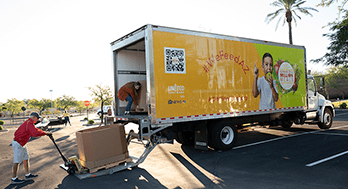 A man in a red shirt operates a pallet jack to load boxes onto a truck with a bright yellow "WeFeedAZ" banner featuring a smiling child.