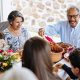 multi-generational family shares a meal outdoors. An older man with glasses serves food from a pot, surrounded by smiling relatives and flowers.