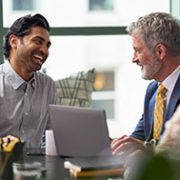 A group of three professionals sit around a table, smiling and engaged in a lively discussion. A laptop and notepad are on the table, suggesting a collaborative meeting.