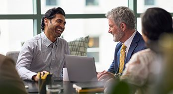 A group of three professionals sit around a table, smiling and engaged in a lively discussion. A laptop and notepad are on the table, suggesting a collaborative meeting.