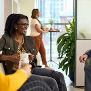Three people sit and talk in an office lounge, each holding a mug, while another person stands by a counter in the background.