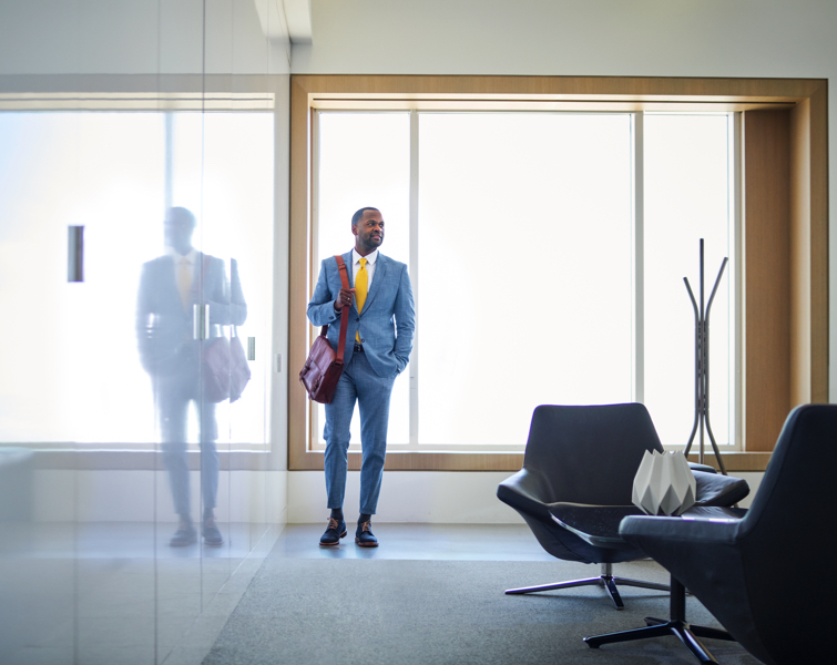 A man in a business suit stands along a reflective wall of glass.