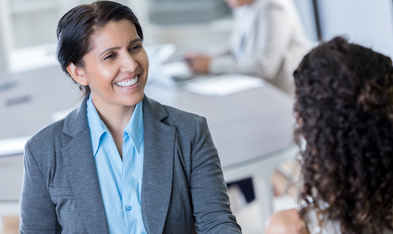 Confident mature Hispanic businesswoman shakes a client or colleague's hand in the office. People are working in the background.