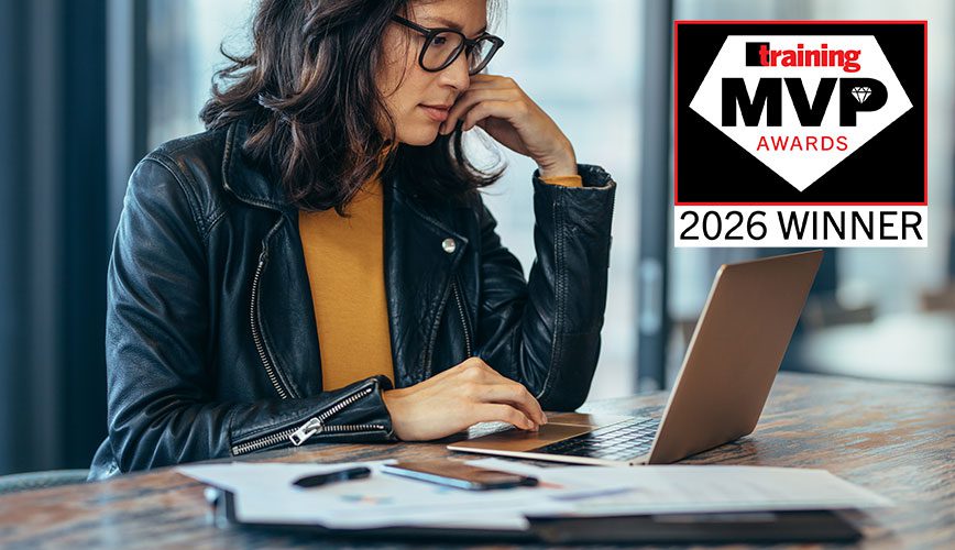 A woman in a black leather jacket and glasses works intently on a laptop at a wooden table, with papers and a pen nearby, in a modern office setting.