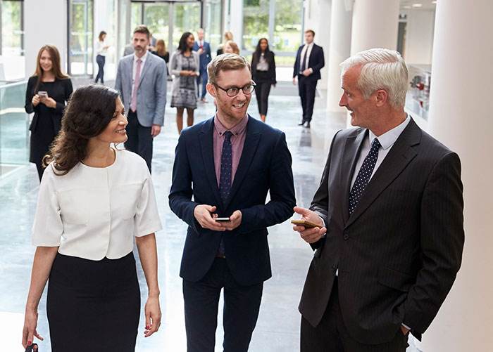 Two men and a women walking and talking in an office building with other employee in the background.