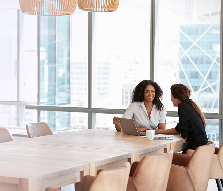 Two women sit across a wooden table in a bright office. One has a laptop, and they are engaged in a friendly conversation, conveying a collaborative tone.