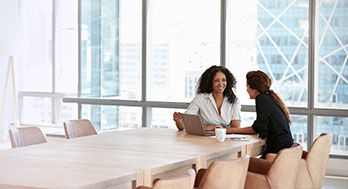 Two women sit across a wooden table in a bright office. One has a laptop, and they are engaged in a friendly conversation, conveying a collaborative tone.