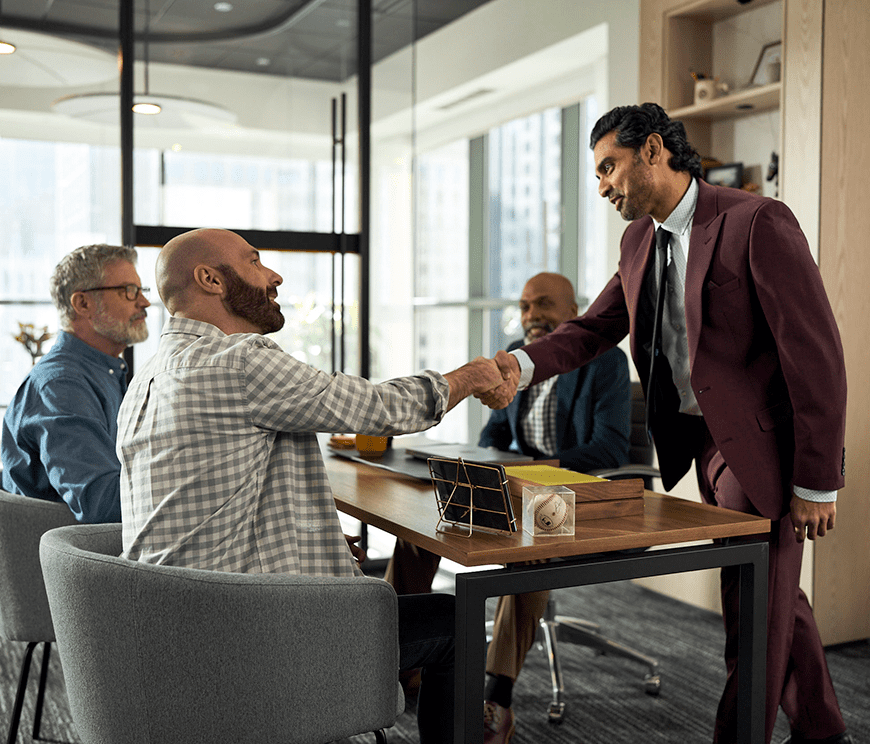 A man in a burgundy suit shakes hands with a seated, bearded man in a plaid shirt at a conference table. Two others men observing.