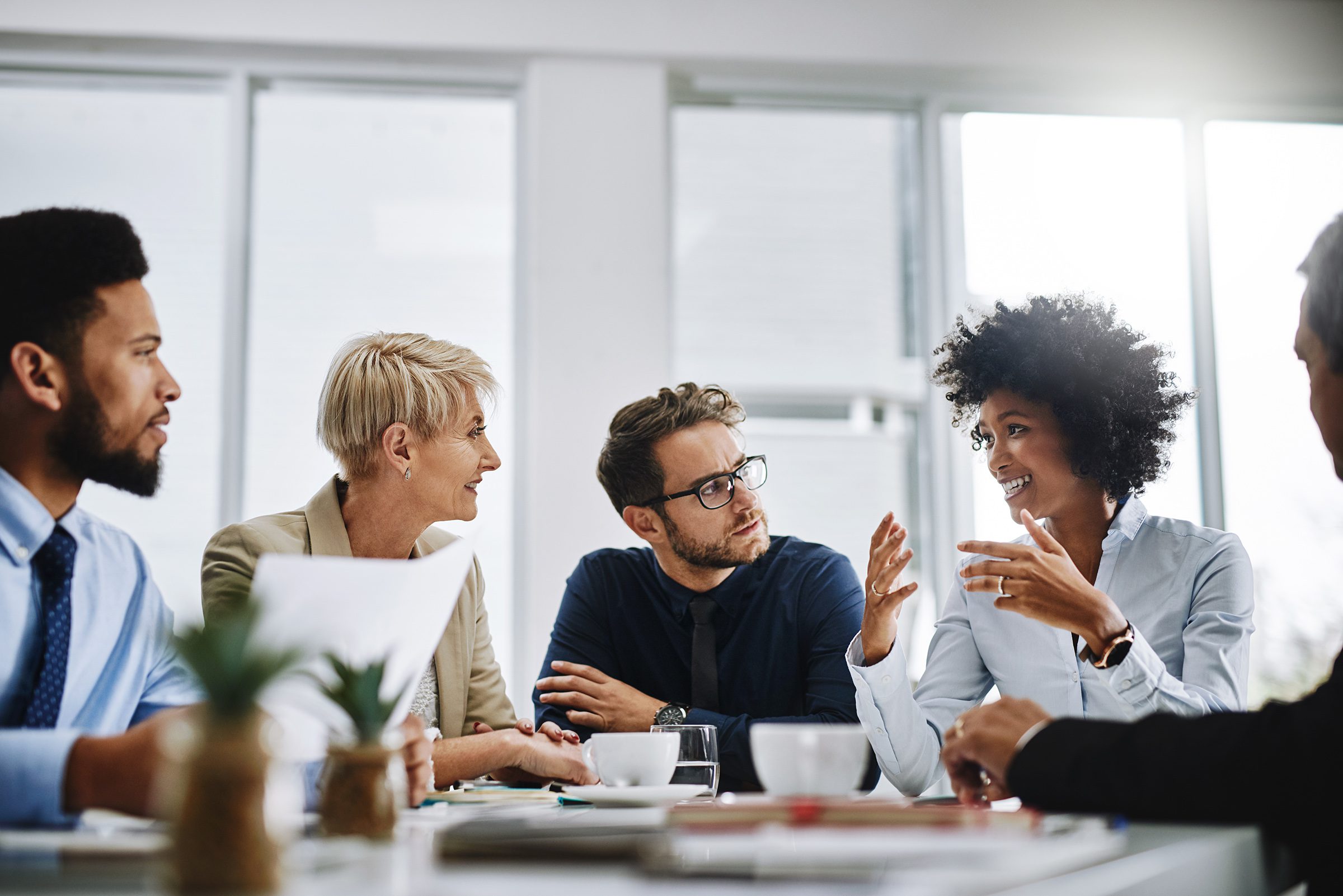 A diverse group of professionals engaged in a lively discussion around a conference table. Bright lighting, plants, and papers create a collaborative atmosphere.