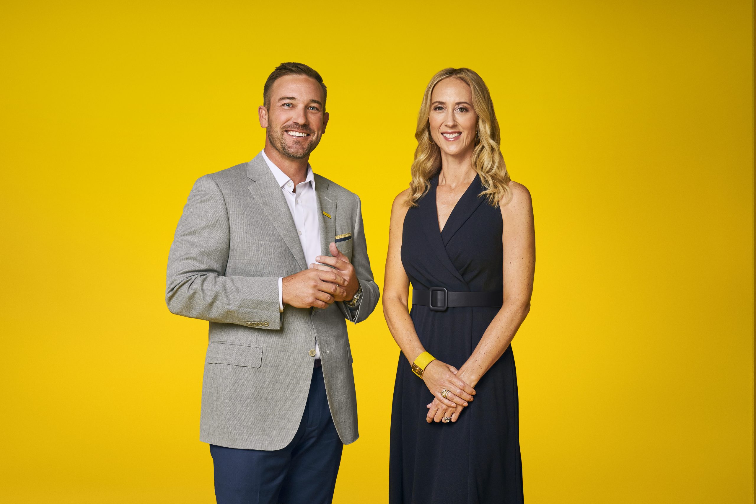 Two professionally dressed colleagues standing side by side and smiling at the camera against a bright yellow background.