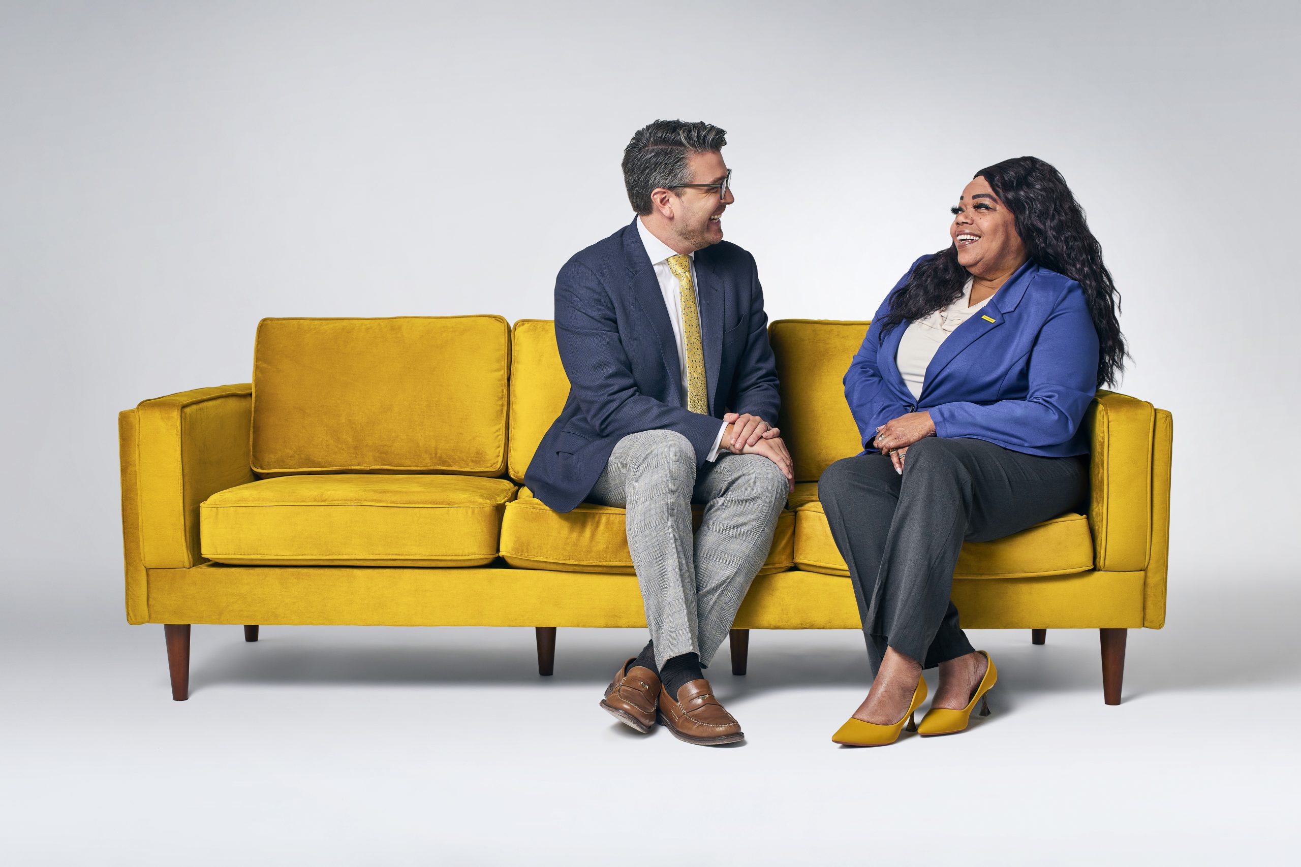 Two professionally dressed colleagues sitting on a bright yellow couch, smiling and engaged in conversation against a light gray background.