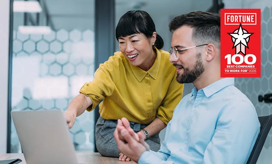 A woman in a yellow shirt smiles and points at a laptop screen with a seated man in glasses. A "Fortune 100 Best Companies to Work For 2026" badge is in the corner.