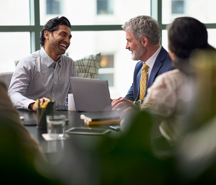 A group of three professionals sit around a table, smiling and engaged in a lively discussion. A laptop and notepad are on the table, suggesting a collaborative meeting.